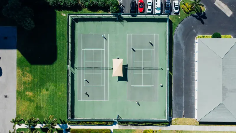 Drone shot capturing a tennis match on dual courts with players and parked cars under clear summer skies