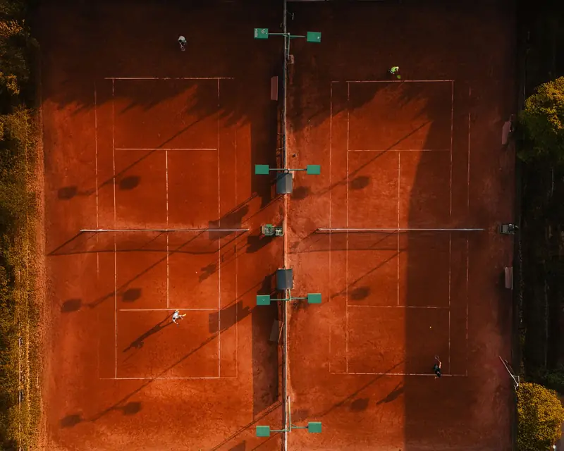 Aerial shot of clay tennis courts with players during golden hour, casting long shadows