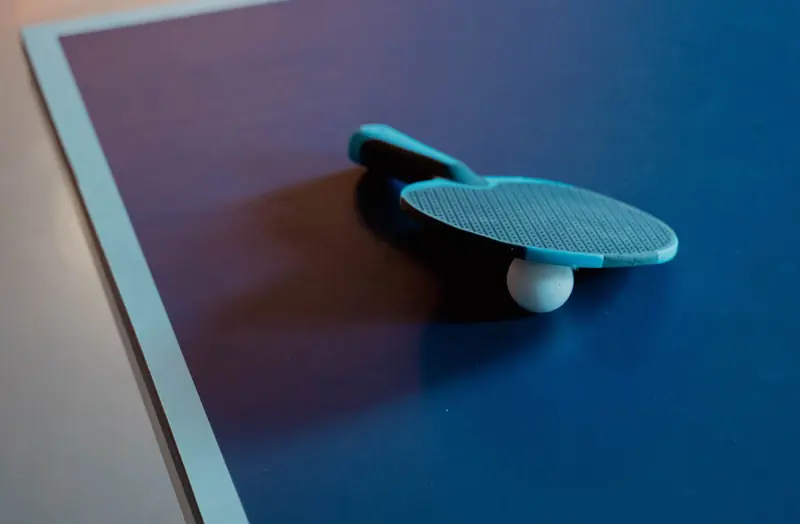 Close-up of a table tennis paddle and ball on a blue table, indoor setting