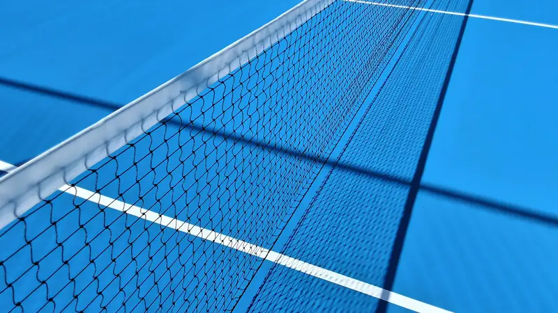 Perspective view of a blue tennis court highlighting the net and shadows under sunlight
