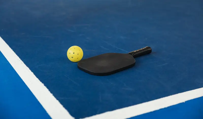 A pickleball paddle and yellow ball on a blue court corner, symbolizing sport and activity