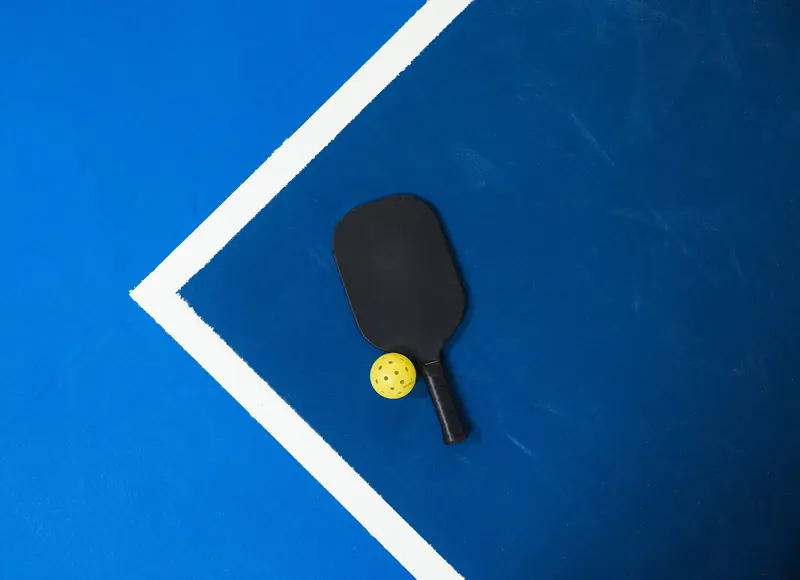 Close-up of a pickleball paddle and ball on a vibrant blue court surface showing geometric lines