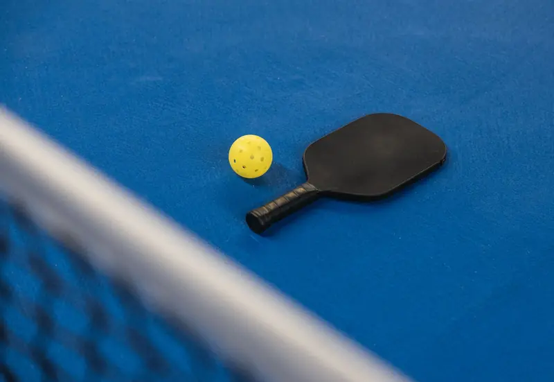 A pickleball paddle and ball resting on a blue court surface with a net in focus